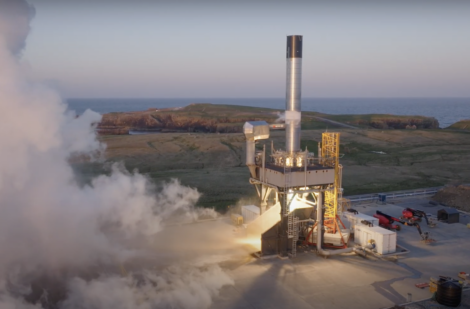 A rocket stands on a launch pad emitting smoke and preparing for liftoff, with a coastal landscape and ocean in the background.