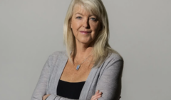 A woman with long blonde hair wearing a gray cardigan and a necklace stands with her arms crossed against a plain background.