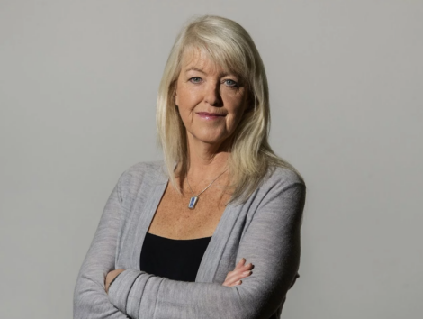 A woman with long blonde hair wearing a gray cardigan and a necklace stands with her arms crossed against a plain background.