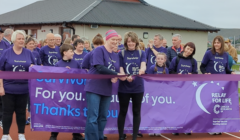 A group of people wearing purple shirts that read "Survivor" stand behind a purple "Relay for Life" banner at an outdoor event. A woman in the center is cutting a ribbon, marking the start of the event.