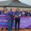 A group of people wearing purple shirts that read "Survivor" stand behind a purple "Relay for Life" banner at an outdoor event. A woman in the center is cutting a ribbon, marking the start of the event.