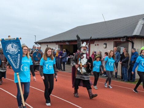 A group of people in blue shirts march beside a costumed person holding a spear and shield during an outdoor event on a running track.