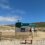 A portable wooden sauna named "Haar Sauna" with a green roof sits on sandy ground, surrounded by a wooden fence. Rolling grassy hills and a clear blue sky are in the background.