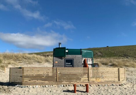 A portable wooden sauna named "Haar Sauna" with a green roof sits on sandy ground, surrounded by a wooden fence. Rolling grassy hills and a clear blue sky are in the background.