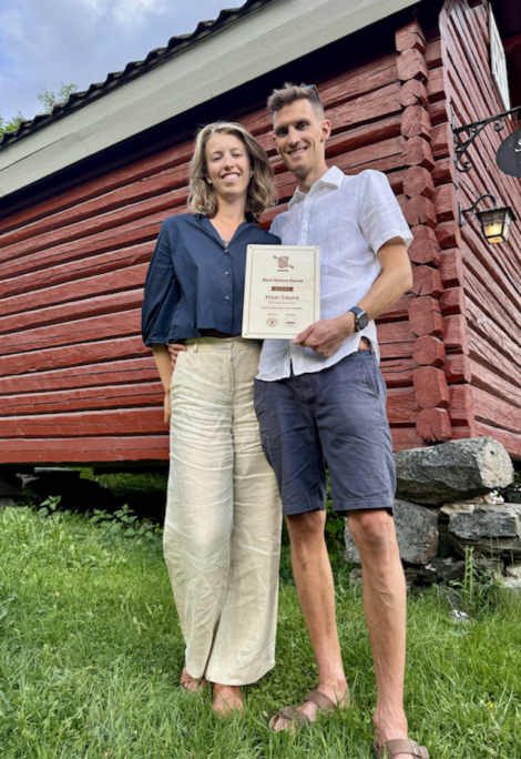 Two people standing on grass in front of a red wooden building, holding a framed certificate.