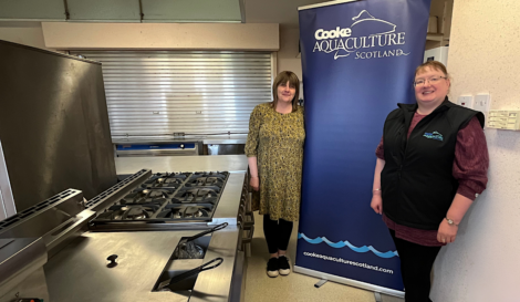 Two women stand in a commercial kitchen beside a banner that reads, "Cooke Aquaculture Scotland." One wears a yellow patterned dress, and the other wears a black vest with the same logo as the banner.