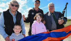 Five people standing outdoors behind a vibrant red, orange, and blue ribbon, with one person holding bagpipes. Two children are also present, along with sunny weather in the background.