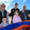 Five people standing outdoors behind a vibrant red, orange, and blue ribbon, with one person holding bagpipes. Two children are also present, along with sunny weather in the background.