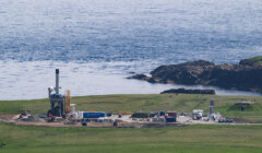 A small coastal rocket launch site with a rocket on the pad, surrounded by support vehicles and trailers, against a backdrop of the ocean and rocky shore.