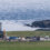 A small coastal rocket launch site with a rocket on the pad, surrounded by support vehicles and trailers, against a backdrop of the ocean and rocky shore.