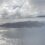 Aerial view of a coastal island with rugged terrain, surrounded by the ocean, under a partly cloudy sky. The edge of an aircraft wing is partially visible on the right side.