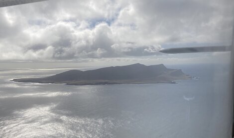 Aerial view of a coastal island with rugged terrain, surrounded by the ocean, under a partly cloudy sky. The edge of an aircraft wing is partially visible on the right side.