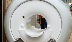 A large mri machine with a white cylindrical body and cables attached, displayed outdoors with people in the background observing.