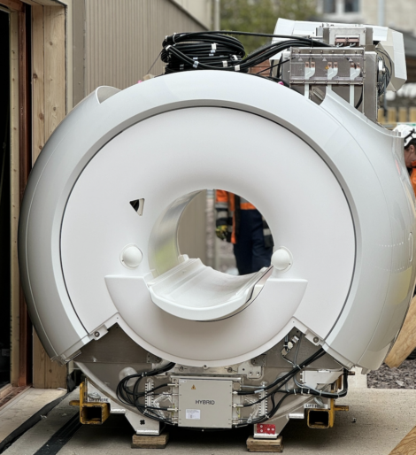 A large mri machine with a white cylindrical body and cables attached, displayed outdoors with people in the background observing.