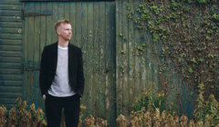 A man in a black blazer and striped shirt standing thoughtfully beside a wooden shed, surrounded by overgrown grass.