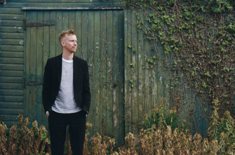 A man in a black blazer and striped shirt standing thoughtfully beside a wooden shed, surrounded by overgrown grass.