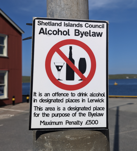 Sign on a pole stating "Shetland Islands Council Alcohol Byelaw." It is an offense to drink alcohol in designated places in Lerwick. Maximum penalty £500. The sign shows a no alcohol symbol.