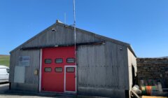 A small corrugated metal building with a red garage door and an antenna on top. A vehicle is partially visible on the left side. The building is on a clear, sunny day.