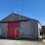 A small corrugated metal building with a red garage door and an antenna on top. A vehicle is partially visible on the left side. The building is on a clear, sunny day.