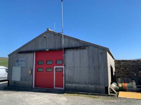 A small corrugated metal building with a red garage door and an antenna on top. A vehicle is partially visible on the left side. The building is on a clear, sunny day.