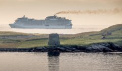 A large cruise ship sails near a coastline with a historic stone structure and ruins visible on the grassy terrain in the foreground.