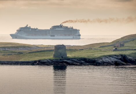 A large cruise ship sails near a coastline with a historic stone structure and ruins visible on the grassy terrain in the foreground.