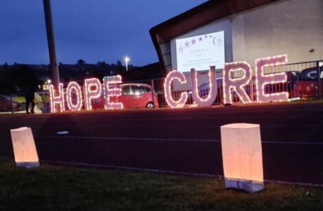 Large, illuminated letters spelling "HOPE" and "CURE" are displayed on a track field at dusk, with a person in the background and paper lanterns in the foreground.