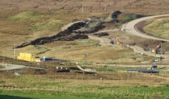 Aerial view of a construction site with earth-moving equipment on rugged terrain, featuring roads, a yellow site office, and workers.