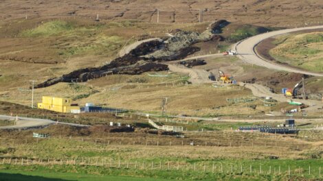 Aerial view of a construction site with earth-moving equipment on rugged terrain, featuring roads, a yellow site office, and workers.