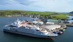 A large cruise ship docked at a port with buildings and smaller boats nearby. Green hills and a village are visible in the background under a partly cloudy sky.