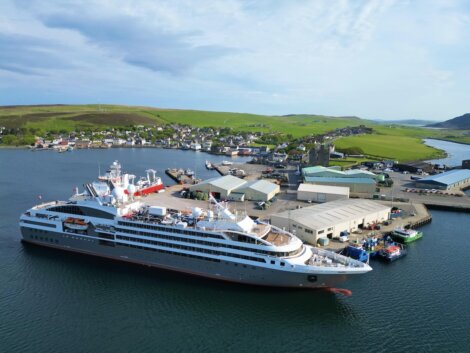 A large cruise ship docked at a port with buildings and smaller boats nearby. Green hills and a village are visible in the background under a partly cloudy sky.