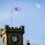 A clock tower with a flag displaying an emblem flies above Lerwick Town Hall. The sky is clear and blue.