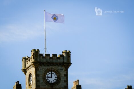 A clock tower with a flag displaying an emblem flies above Lerwick Town Hall. The sky is clear and blue.