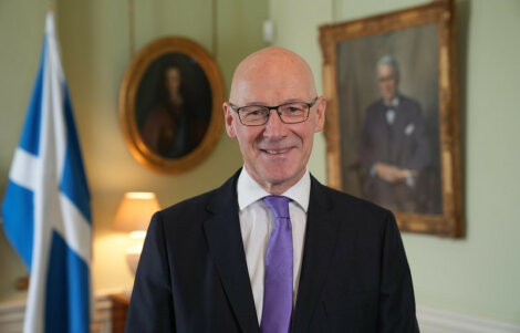 A bald man in a suit and purple tie stands in front of a wall with portraits and a Scottish flag to his side.