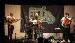 Three musicians performing at the shetland folk festival, featuring a guitarist, a drummer, and an accordion player on stage.