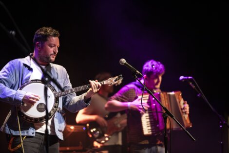 A banjo player and an accordionist perform on stage, illuminated by soft stage lighting with a guitarist in the background.