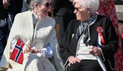 Two elderly women sit and talk outdoors. One holds a Norwegian flag and wears a cream shawl, while the other wears a black jacket with a red, white, and blue ribbon.