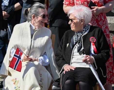 Two elderly women sit and talk outdoors. One holds a Norwegian flag and wears a cream shawl, while the other wears a black jacket with a red, white, and blue ribbon.