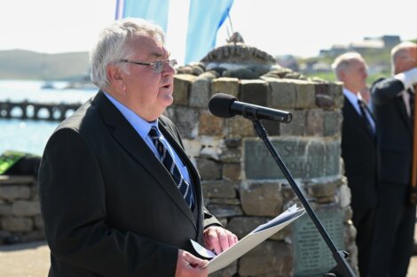 A man in a suit speaks into a microphone while holding papers at an outdoor event. In the background, there is a stone structure and several other people.