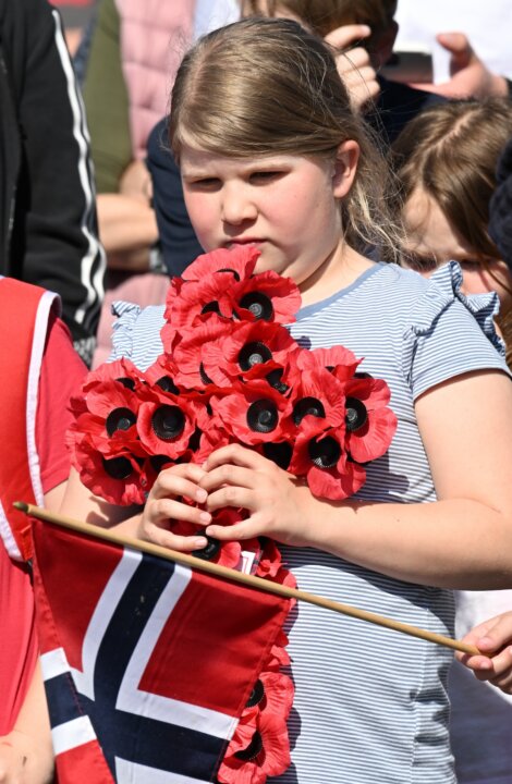 A young girl holds a wreath made of red poppies and a Norwegian flag, standing outdoors among other people.