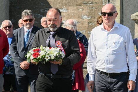 A group of men stand together, one in the front holding a floral wreath with red, white, and blue flowers. They appear to be in an outdoor, possibly ceremonial, setting.