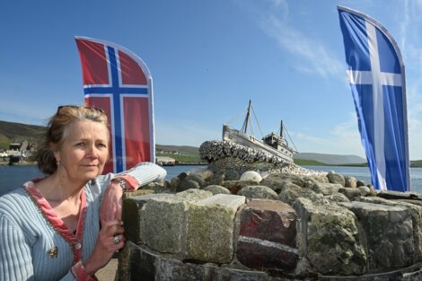 A woman stands next to a stone structure with model boat, flanked by Norwegian and Scottish flags in a scenic outdoor location.