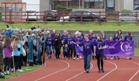 A group of people in purple shirts walk on a track holding a banner that reads "Relay for Life." Spectators, some in medieval costumes, stand nearby.