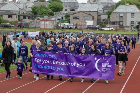 A large group of people, many wearing purple "Survivor" shirts, walk on a track holding a "Relay For Life" banner with the message "Survivors. For you. Because of you. Thanks to you.