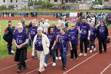 A group of people wearing purple "Survivor" t-shirts walk on a track at an outdoor event, with others in the background.