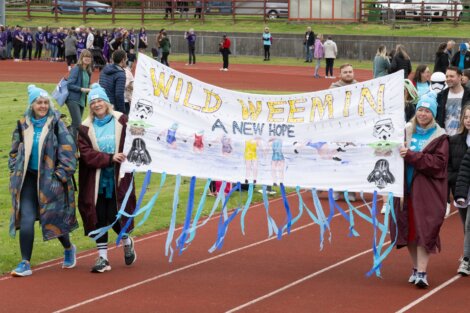 A group of people walk on a track carrying a banner that reads, "WILD WEEMIN: A NEW HOPE" with Star Wars characters and decorations. Other people are visible in the background on the track.