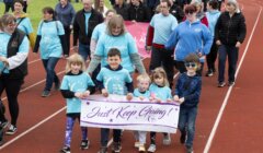 A group of people, including children and adults, walk on a track in matching blue shirts. The children in front hold a banner reading "Just Keep Going!.