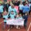 A group of people, including children and adults, walk on a track in matching blue shirts. The children in front hold a banner reading "Just Keep Going!.