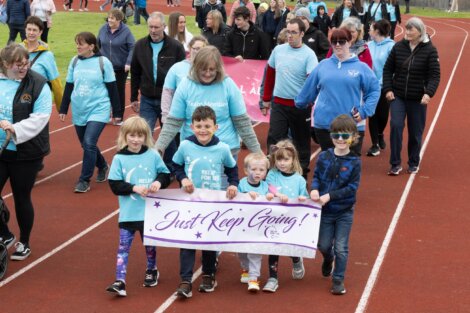 A group of people, including children and adults, walk on a track in matching blue shirts. The children in front hold a banner reading "Just Keep Going!.