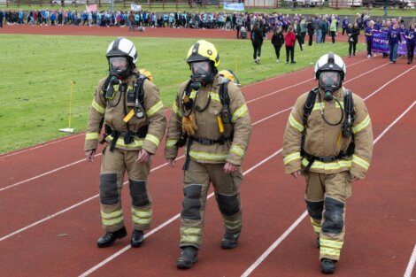 Three firefighters in full gear, including helmets and breathing apparatus, walk side-by-side on a track. A crowd follows behind them in the background.